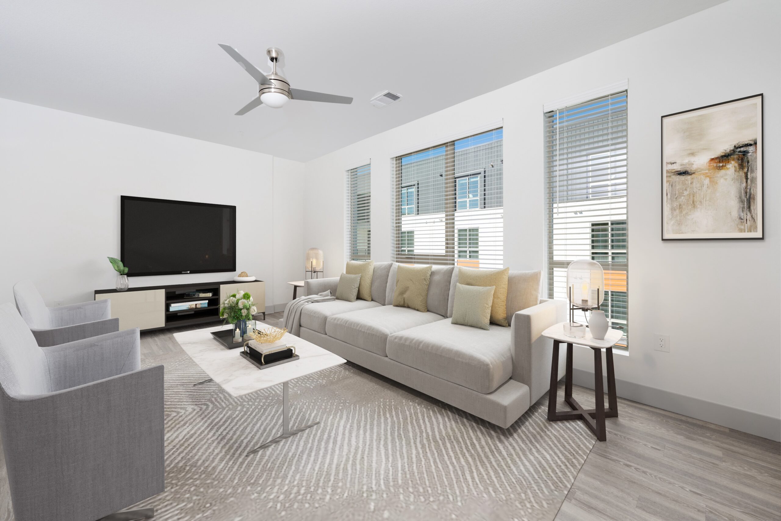 A neutral living room with a large, light gray sofa and two gray armchairs centered around a white marble coffee table. The room has a ceiling fan, a TV on a low console, a patterned area rug, and several windows with blinds overlooking an apartment building exterior.